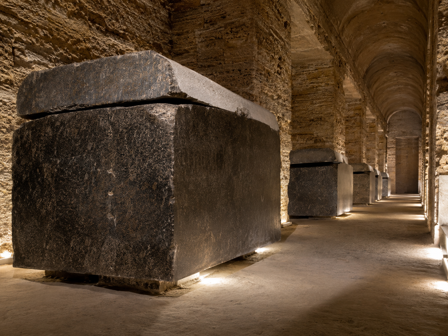 AI-generated illustrative reconstruction of the Serapeum's main underground gallery, showing a long stone corridor lined with dark granite sarcophagi in side niches.