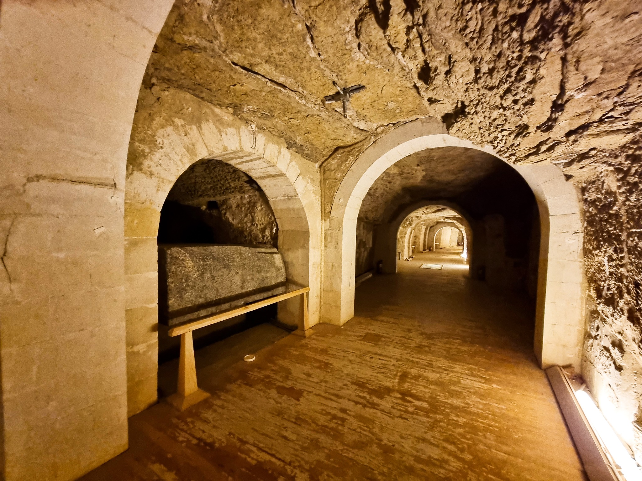 Photograph looking down a vaulted stone gallery of the Serapeum of Saqqara, with a granite sarcophagus visible in a side niche and further arched chambers receding into the distance.